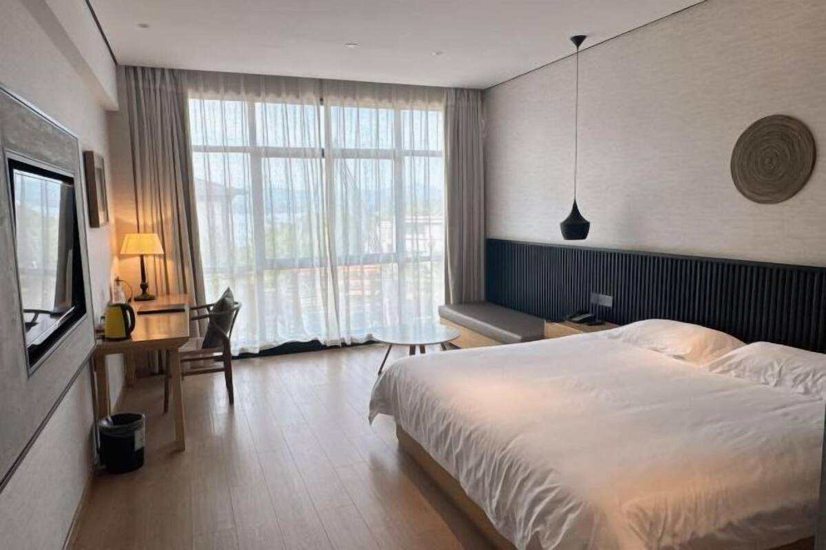 Une chambre d’hôtel avec un grand lit, un bureau avec une chaise, une fenêtre avec des rideaux et un luminaire fixé au mur.