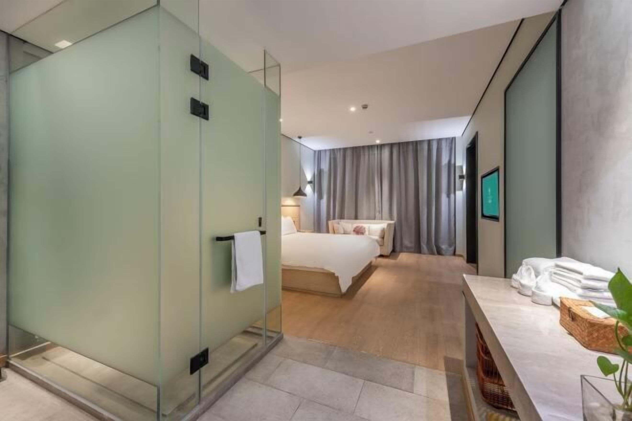 A modern hotel room with a glass partition, a bed, a bathroom with a vanity, and a view of the city.