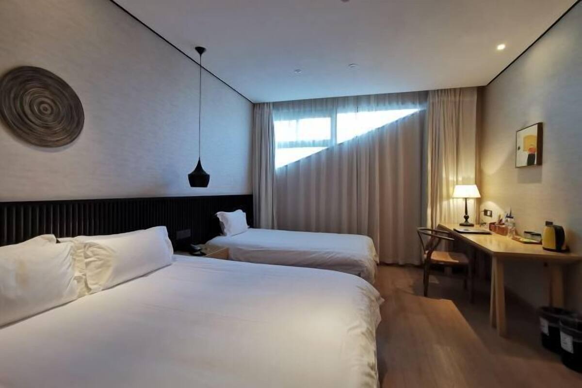 Une chambre d’hôtel avec deux lits, un bureau, une chaise, une lampe et une fenêtre avec des rideaux.