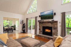 A spacious living room with a stone fireplace, a television mounted above it, and a large window overlooking a deck.