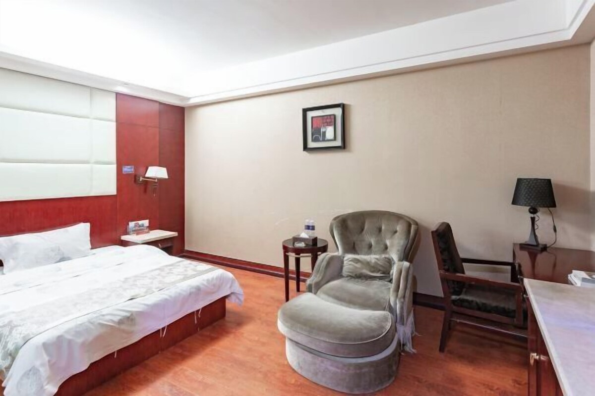 A hotel room with a bed, a grey armchair, a small table with a lamp, and a framed picture on the wall.