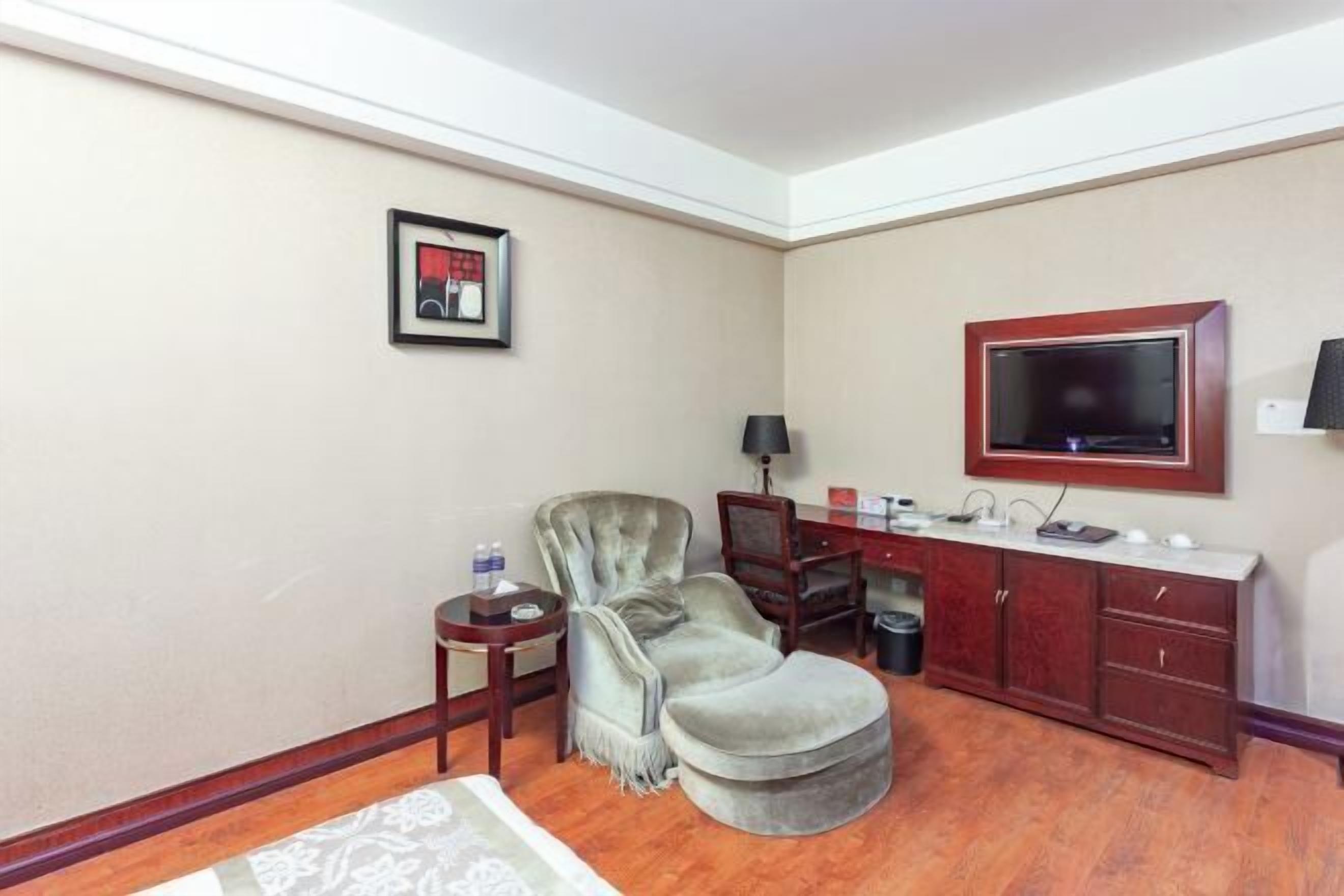 A room with a grey armchair, a small table with a lamp, a TV on the wall, and a wooden cabinet.