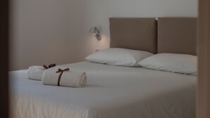 A neatly made bed with white linens and two rolled towels with brown ribbons, a wall-mounted lamp, and a plain wall. - WeLive Trapani - Superior Second Floor Apartment (Trapani)