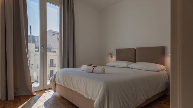 A bedroom with a large bed, a view through a sliding door, and a nightstand with a lamp. - WeLive Trapani - Superior Second Floor Apartment (Trapani)