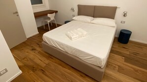 Una cama bien tendida con sábanas blancas y toallas enrolladas, un escritorio pequeño con una silla, y una ventana con cortina.