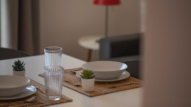Mesa para comer con cubertería de vidrio transparente, platos y pequeñas macetas, con una lámpara roja al fondo.