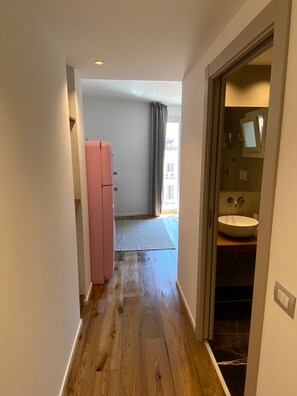 Un couloir étroit avec un plancher en bois, un réfrigérateur rose et une salle de bain équipée d’un lavabo et d’un miroir.