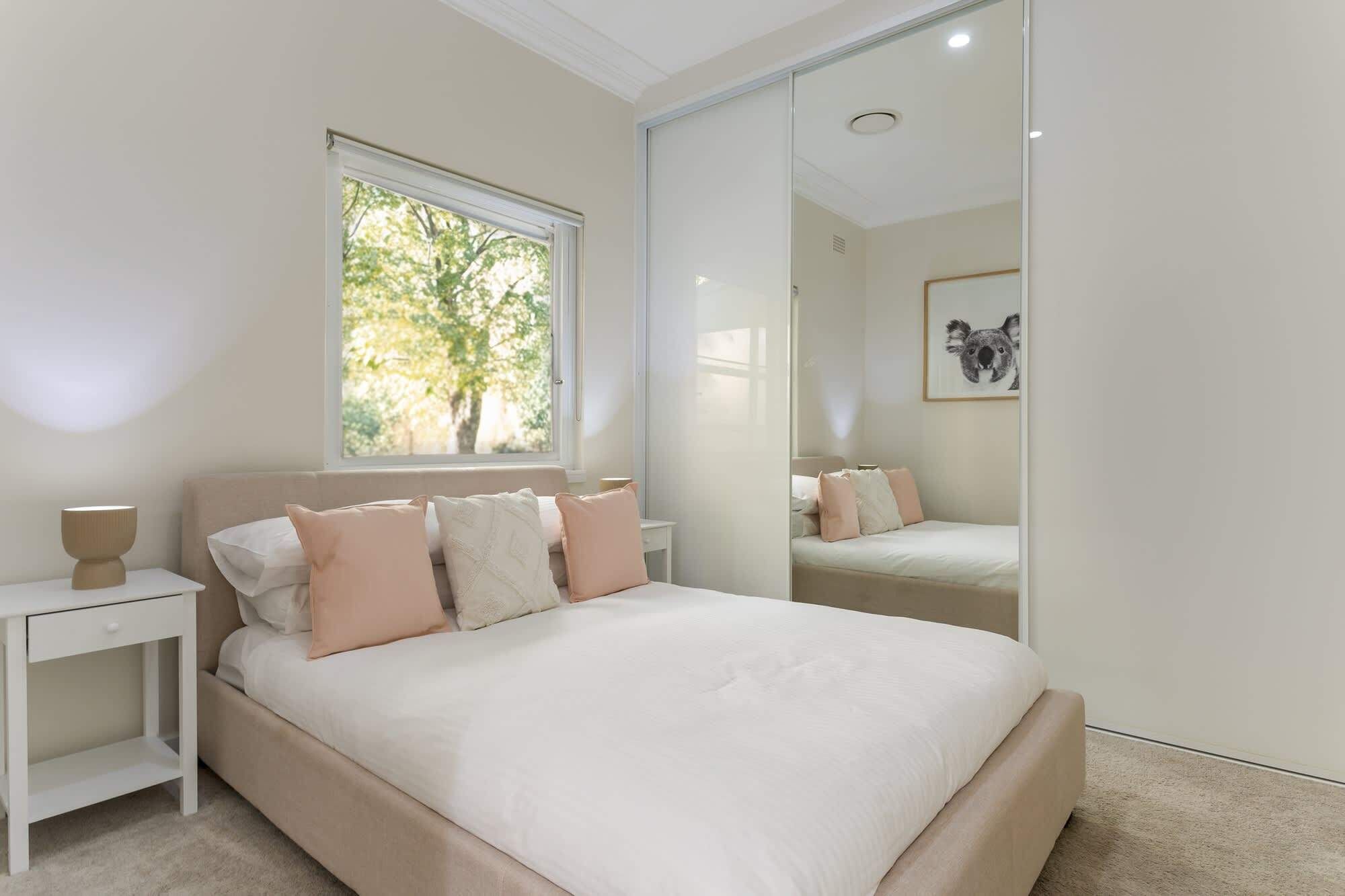 A bedroom with a large bed, a bedside table, a window with a view of trees, and a mirror reflecting the bed.