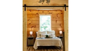 Dormitorio de cabaña de madera con una cama, dos mesitas de noche, una ventana con vistas y un recorte con forma de oso en el techo.