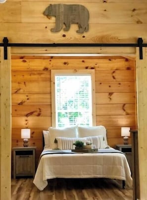 A wooden cabin bedroom with a bed, two bedside tables, a window, and a bear decoration on the wall. - NEW! COZY 4 Guest Cabin in Damascus! PET FRIENDLY! (Damascus)
