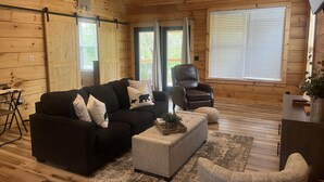 Un interior de cabaña acogedor con un sofá negro, un reposapiés gris, un sillón de cuero y piso de madera.