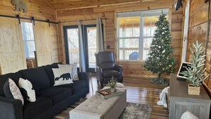 Una sala de estar acogedora tipo cabaña con un sofá de color oscuro, un reposapiés gris y una mesa de centro de madera. Se ve un árbol de Navidad al fondo, y una pequeña mesita de noche con una planta.