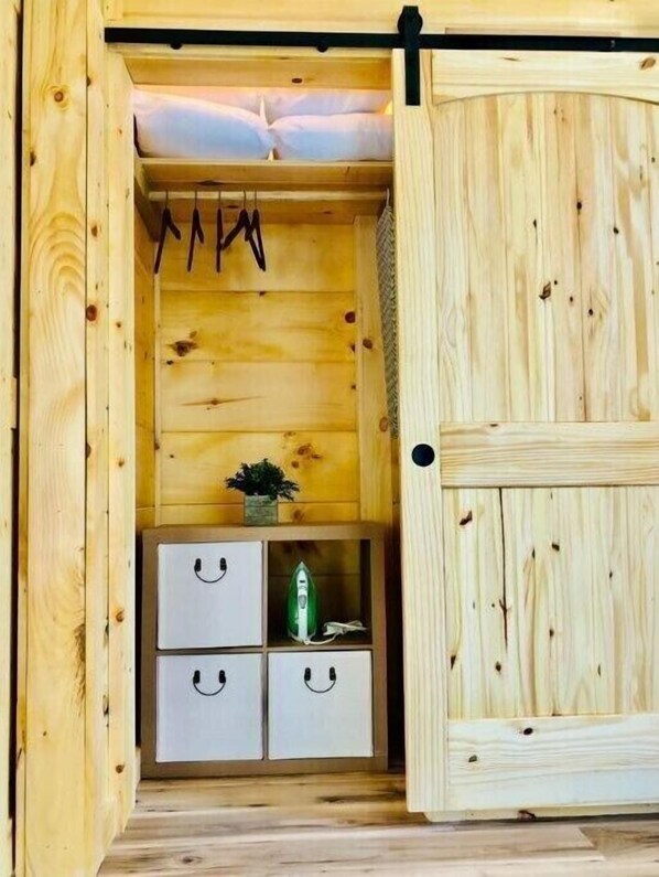 Interior de una cabaña de madera con una cama, un perchero, una planta pequeña y cajas de almacenamiento.