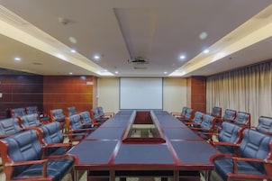Un salón de conferencias con una larga mesa y filas de sillas, una pantalla de proyector y un amplio ventanal con cortinas.