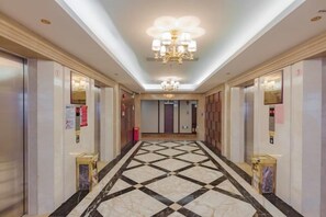 Un couloir d’hôtel avec un sol à motifs géométriques, des ascenseurs et un lustre.