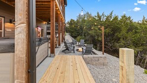 Eine Holzterrasse mit Sitzgelegenheiten im Freien und einer Feuerstelle, umgeben von Bäumen und wolkenlosem Himmel.
