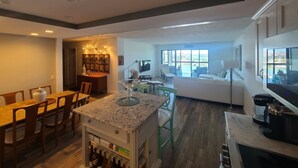 A modern kitchen with a dining area, a living room with a sofa and a TV, and a view of a pool.