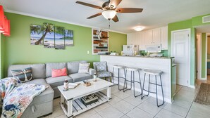 A compact living space with a kitchenette, a ceiling fan, a sofa, a coffee table, and a bar stool area. - Pool Views, Good Times and Beach Vibes at Sea Breeze! (Galveston)