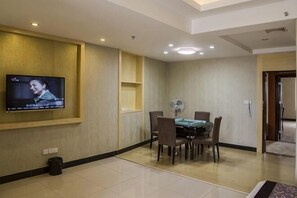 Ein Hotelzimmer mit an der Wand montiertem Fernseher, einem Esstisch mit Stühlen, einem Ventilator und einer Tür zu einem weiteren Raum.