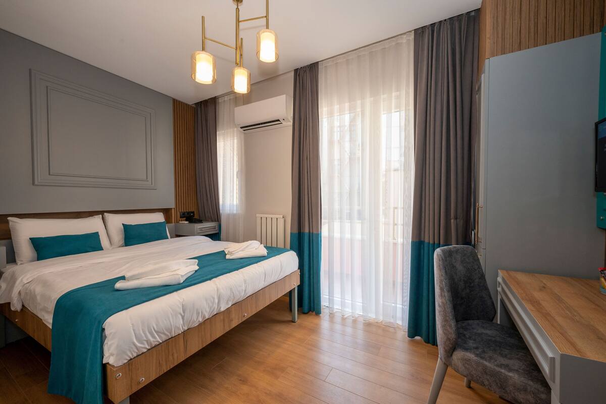 A hotel room with a wooden bed, a desk, and a chair. There is a window with curtains, an air conditioning unit, and a television on the wall.