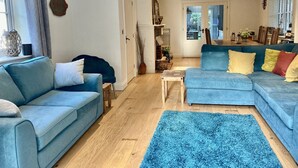 Ein geräumiges Wohnzimmer mit einem blauen Sofa, einem passenden blauen Sessel und einem blauen Teppich. Ein Holztisch und ein kleiner Beistelltisch gehören zur Einrichtung. Der Raum verfügt über Holzdielen und ein großes Fenster mit Vorhängen.