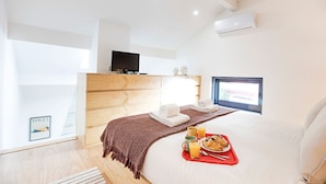 Et moderne hotelværelse med en trækommode, et fladskærms-TV, en stor seng med et brunt tæppe, to håndklæder, et morgenmadsserveringsfad med croissanter og appelsinjuice, samt et vindue med udsigt.