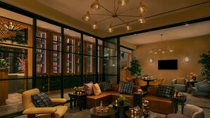 A modern living room with a sofa, armchairs, and coffee tables. There are shelves with plants and a TV mounted on the wall. - Moxy Brooklyn Williamsburg (Brooklyn)