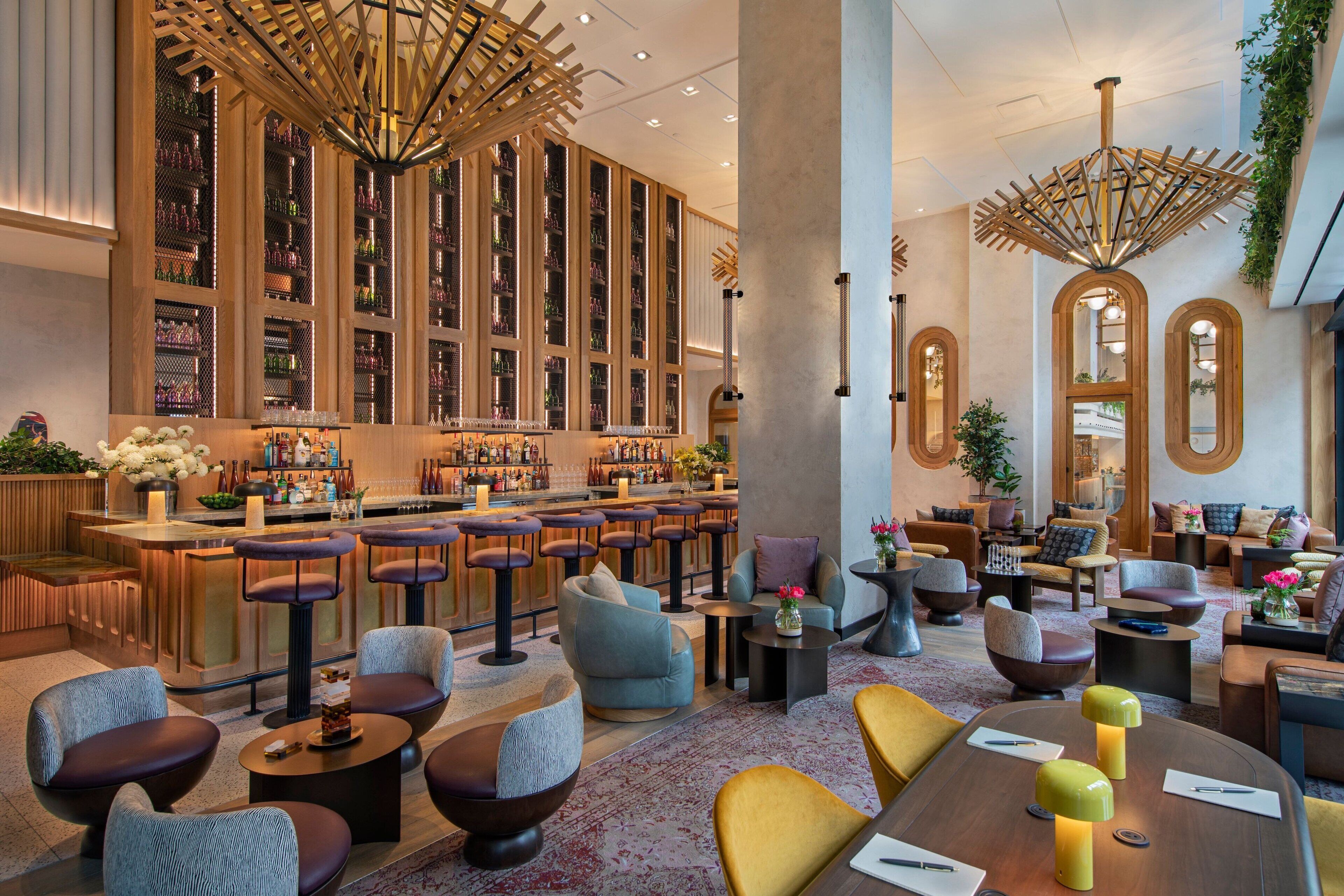 Zona de bar moderna con estanterías altas llenas de botellas, diversas opciones de asientos que incluyen taburetes y sillas acolchadas, y una barra central.