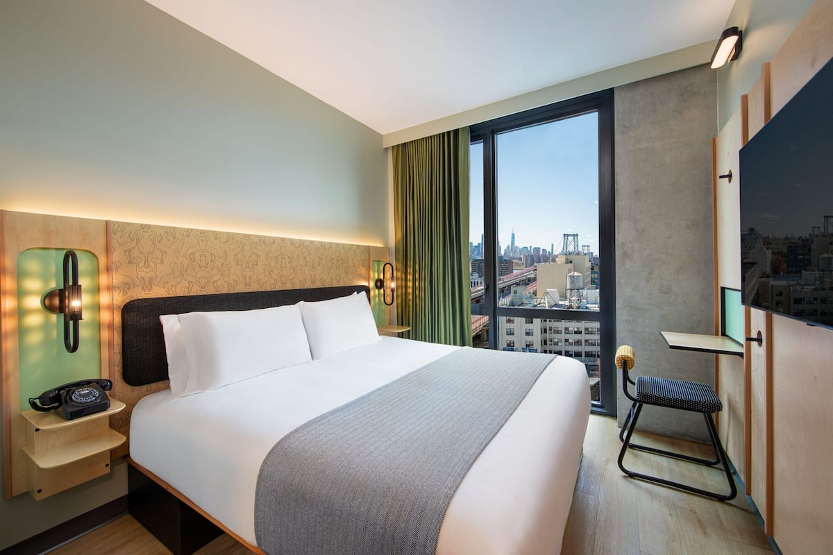A modern hotel room with a large bed, a desk, a chair, and a city view.