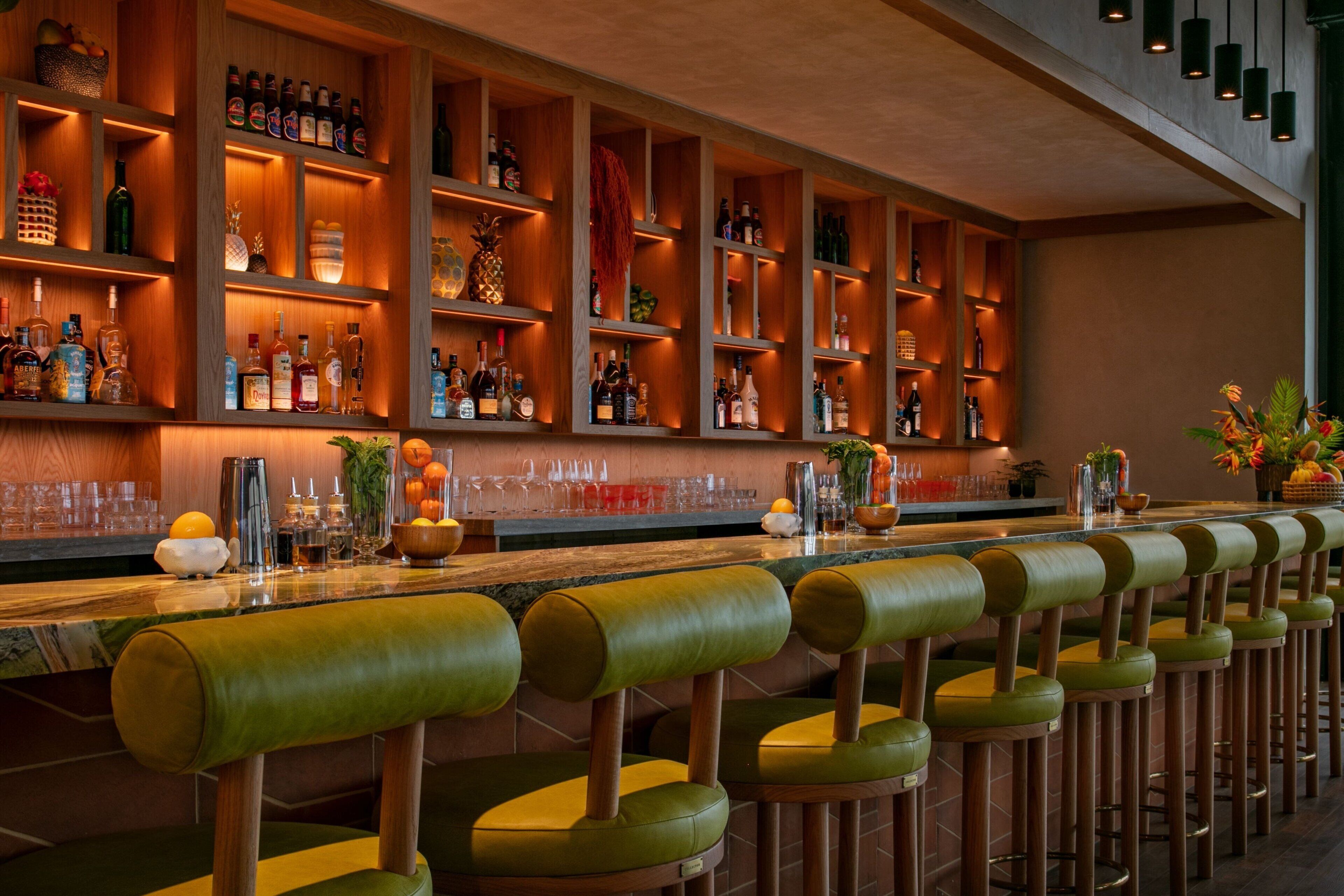 Zona de bar con estantes de madera llenos de botellas, una barra larga con taburetes y objetos decorativos.