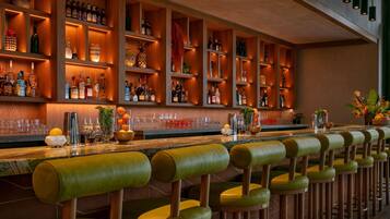 Zona de bar con estantes de madera llenos de botellas, una barra larga con taburetes y objetos decorativos.