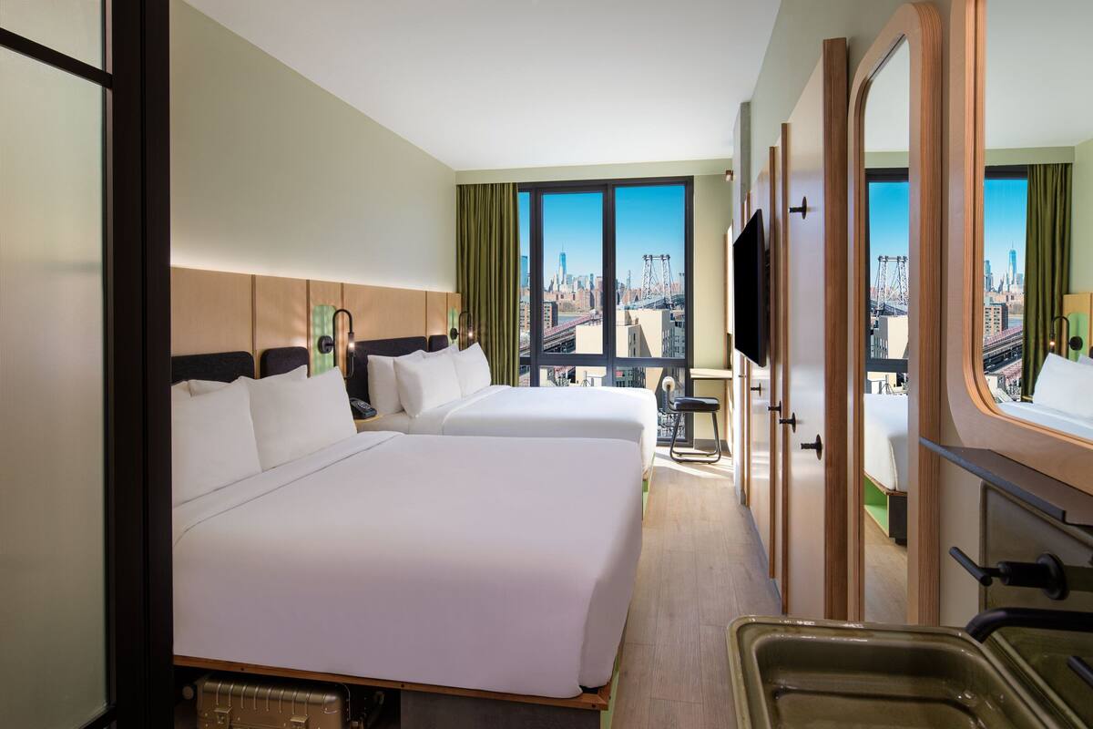 A hotel room with two beds, a large window with a city view, a TV, and a bathroom with a mirror and sink.