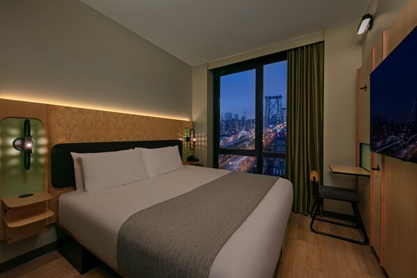 Habitación de hotel con una cama grande, un escritorio, una silla y vistas de la ciudad de noche.