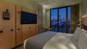 A hotel room with a large bed, a city view through the window, a wooden wardrobe, and a flat-screen TV on the wall.