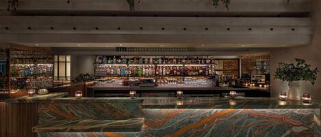 Una zona de bar con mostrador de mármol, estanterías llenas de botellas de licor y plantas colgantes.