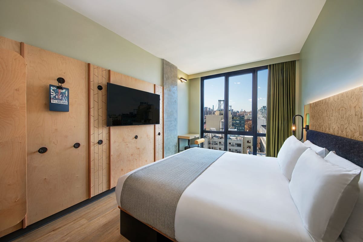 A hotel room with a large bed, a TV mounted on a wooden panel wall, a city view through large windows, and a small table with a chair.