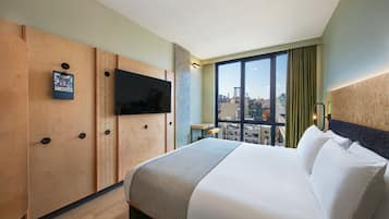 A hotel room with a large bed, a TV mounted on a wooden panel wall, a city view through large windows, and a small table with a chair.