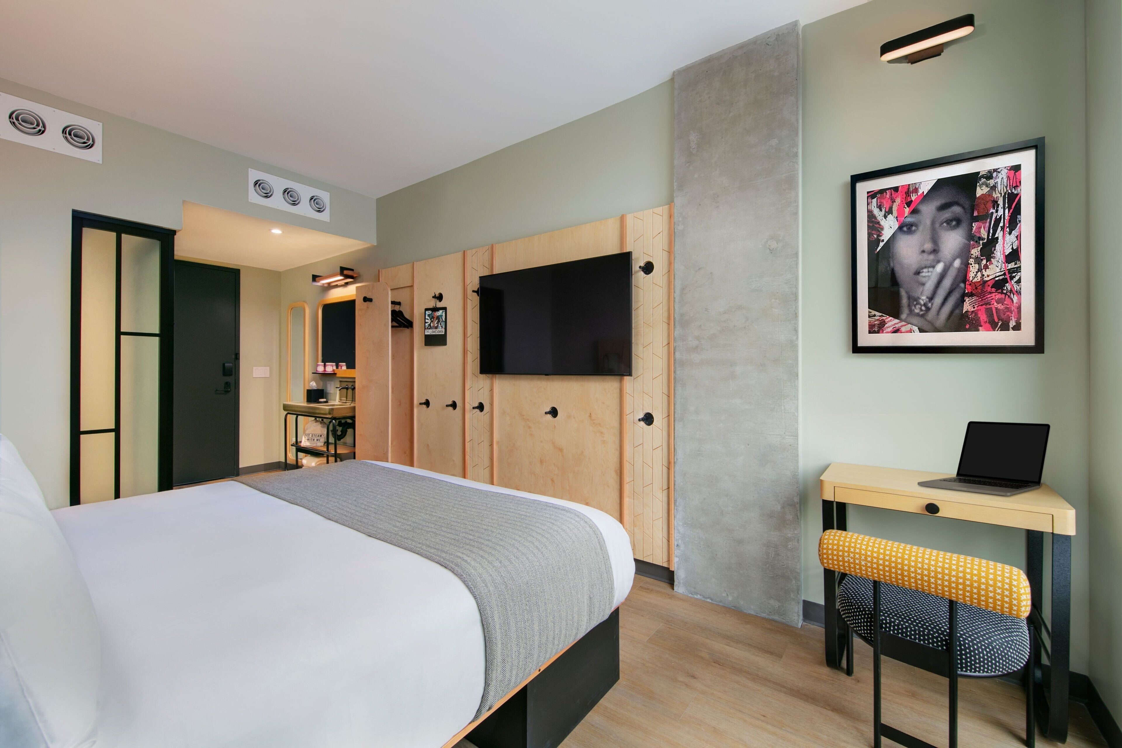 A modern hotel room with a large bed, a desk with a laptop, a TV mounted on the wall, and a wardrobe with a mirror.