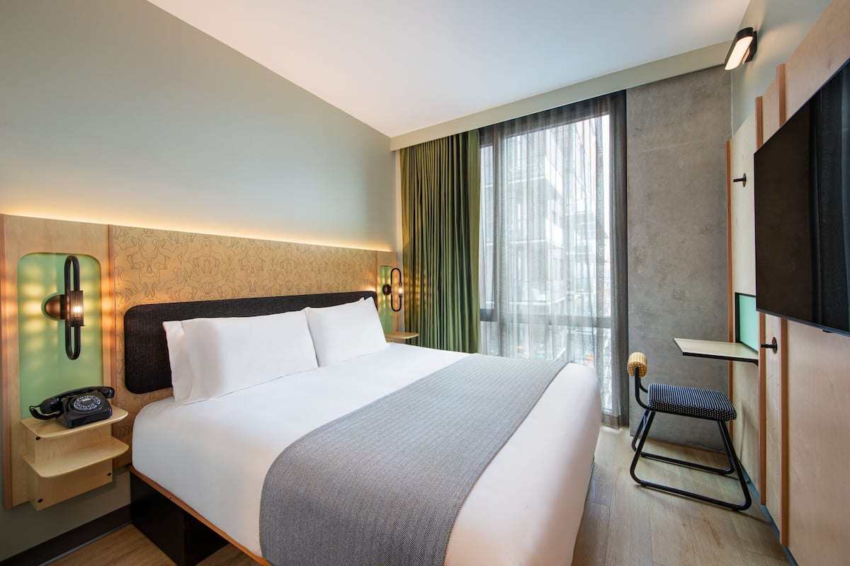 A hotel room with a large bed, a nightstand with a telephone, a small stool, and a TV mounted on the wall.