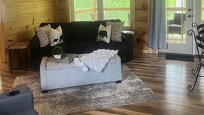 Indbydende interiør i en hytte med træloft, en sofa med dekorative puder, et lille bord med en plante og udsigt gennem et stort vindue.