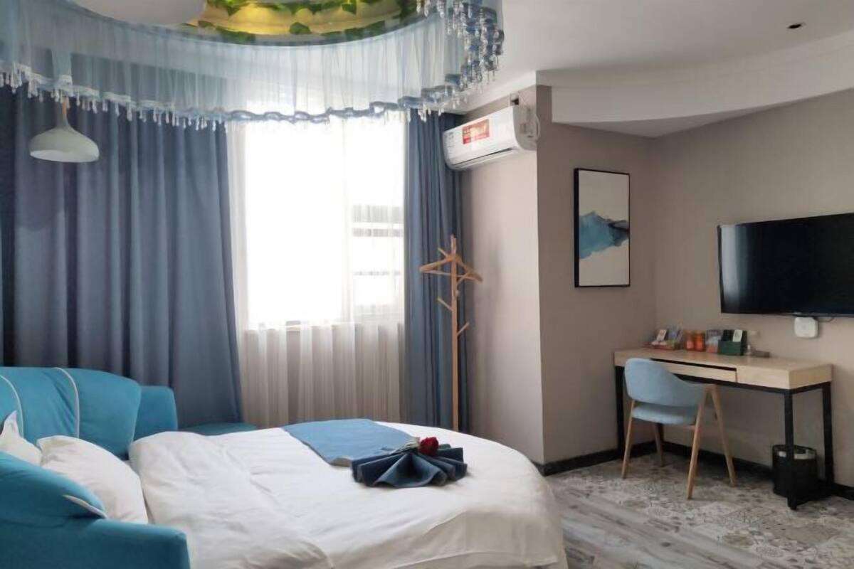 Habitación de hotel con cama, escritorio, silla, televisor y ventana con cortinas.