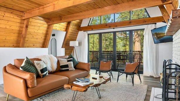 Un salon chaleureux avec un canapé en cuir marron, une table basse et des fauteuils en osier. Une grande fenêtre offre une vue sur les arbres, et il y a un balcon.