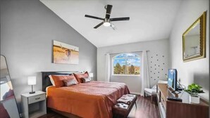 A bedroom with a bed, bedside table, nightstand, ceiling fan, window, and a dresser with a TV. - Cozy Cowboy Condo (Paradise Valley)