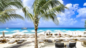 A beach with lounge chairs and umbrellas, a palm tree, and boats in the background. - Fourteen at Mullet Bay (Lowlands)