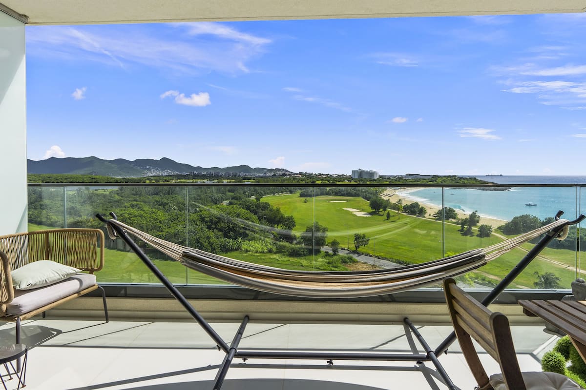Un balcon doté d’un hamac, d’une chaise et d’une table, offrant une vue sur un paysage côtier comprenant un terrain de golf et une plage.