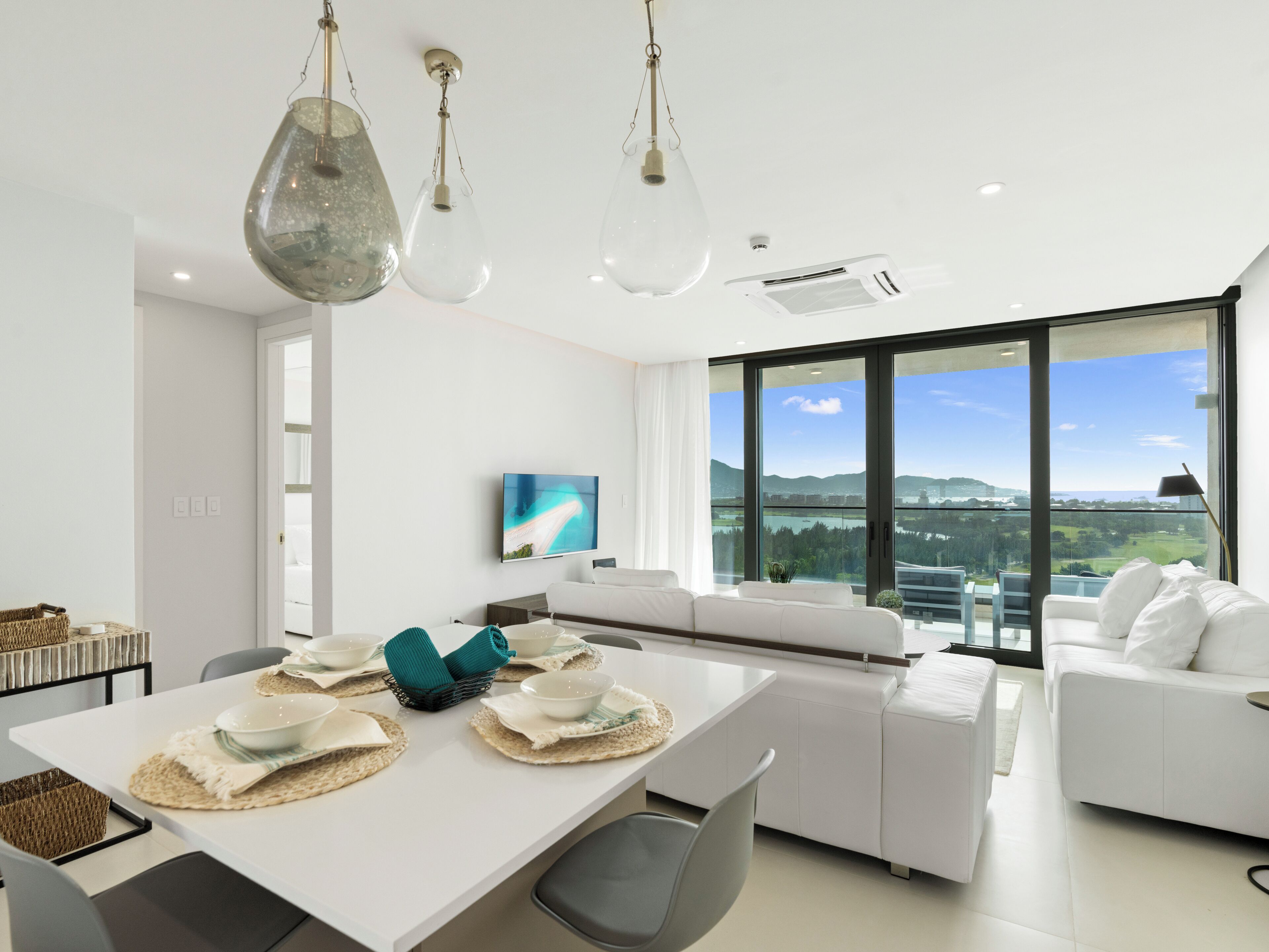 A modern dining area with a white table set for four, a large window with a city view, and a white sofa in the background.