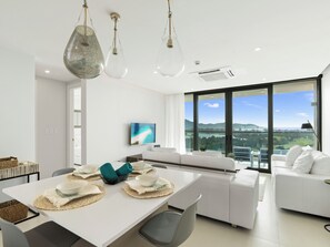 Un espace repas moderne avec une table blanche dressée pour quatre personnes, une grande fenêtre offrant une vue sur la ville et un canapé blanc en arrière-plan.