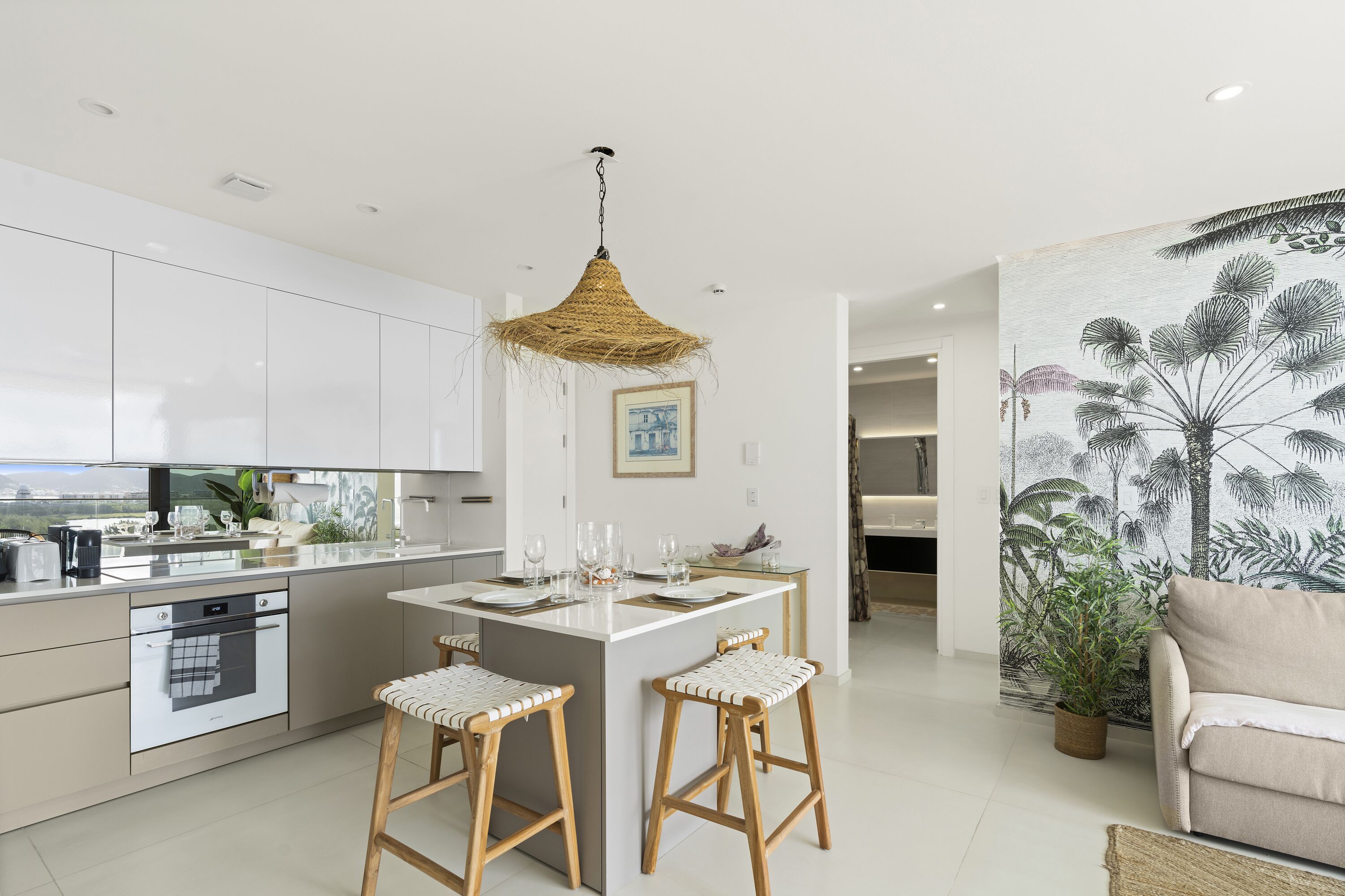 A modern kitchen with white cabinets, a central island with stools, a hanging light fixture, and a tropical-themed wall mural.