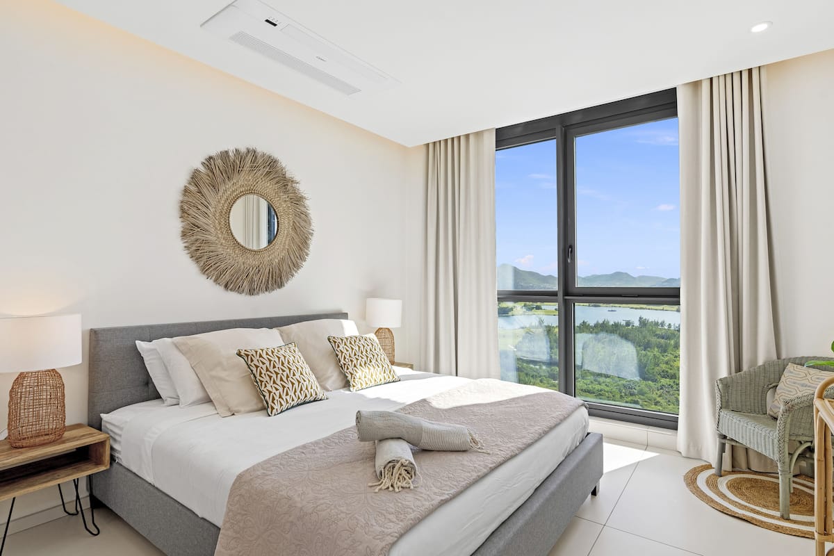 Une chambre à coucher moderne avec un grand lit, un miroir rond et une vue sur un lac, visible à travers une grande fenêtre.