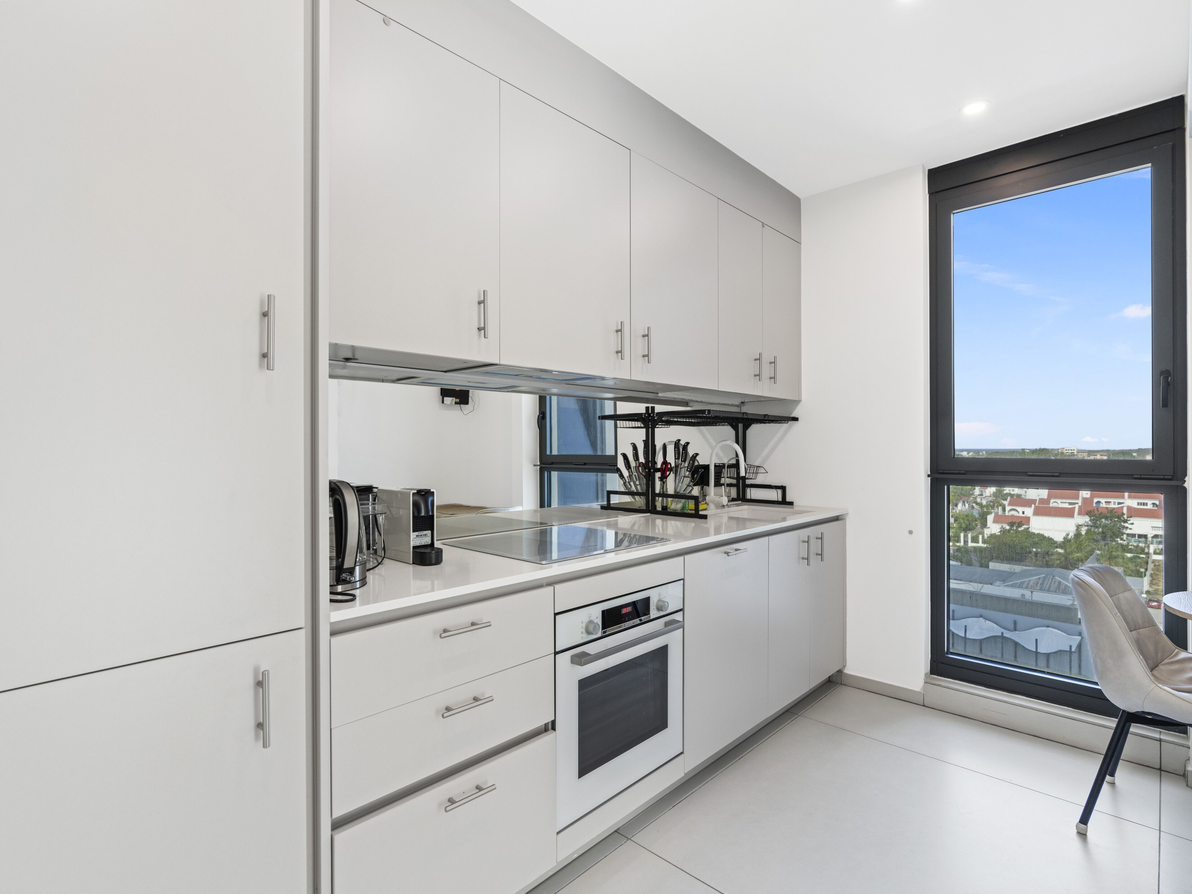 A modern kitchen with white cabinets, a built-in oven, and a large window offering a city view.